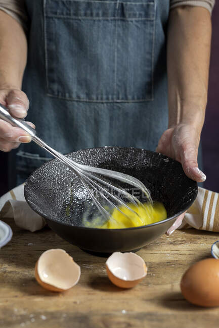 Crop mujer anónima en delantal batiendo huevos en tazón negro en la mesa de madera con limón, harina, mantequilla y palitos de canela ingredientes para pastel - foto de stock