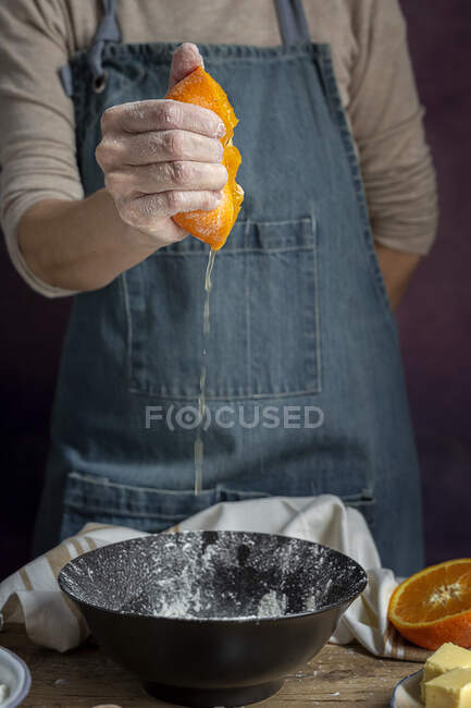 Рука неузнаваемой женщины в фартуке сжимает свежий сочный апельсин над миской, пока готовит тесто за столом. — стоковое фото
