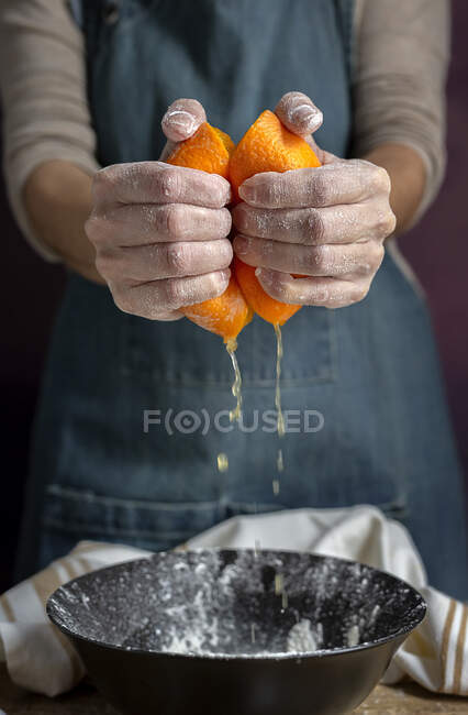 Рука неузнаваемой женщины в фартуке сжимает свежий сочный апельсин над миской, пока готовит тесто за столом. — стоковое фото
