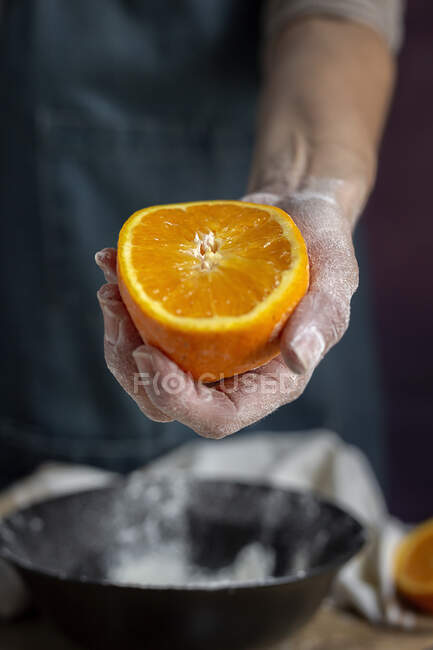 Обрізати руку невпізнаваної жінки, покритої борошном, тримаючи і показуючи камеру свіжий напіврізаний апельсин над мискою, готуючи тісто за столом — стокове фото