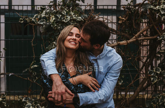 Contenidos joven hombre y mujer abrazándose y besándose de pie cerca de la valla de metal en la calle de la ciudad y disfrutando del día de verano juntos - foto de stock