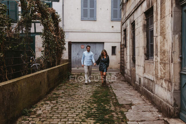 Alegre pareja joven en ropa casual elegante cogidas de la mano y sonriendo mientras camina por la antigua calle estrecha de la ciudad - foto de stock
