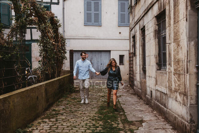 Fröhliches junges Paar in stylischer Freizeitkleidung, Händchen haltend und lächelnd, während es durch die alte enge Straße in der Stadt spaziert — Stockfoto