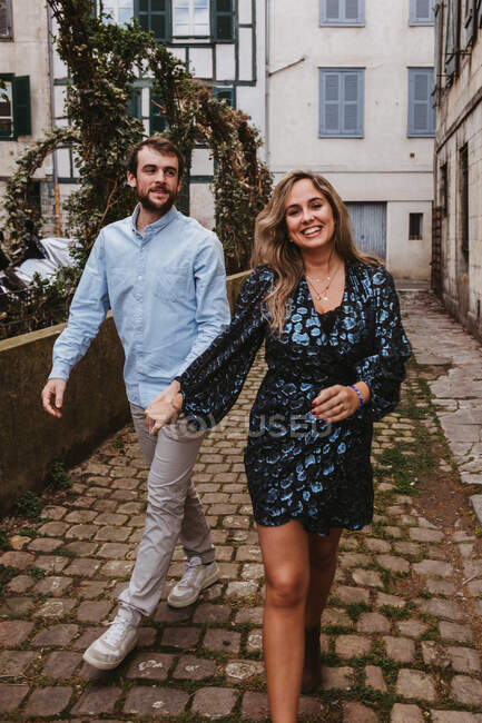 Весела молода пара в стильному повсякденному одязі тримає руки і посміхається під час прогулянки по старій вузькій вулиці міста — стокове фото