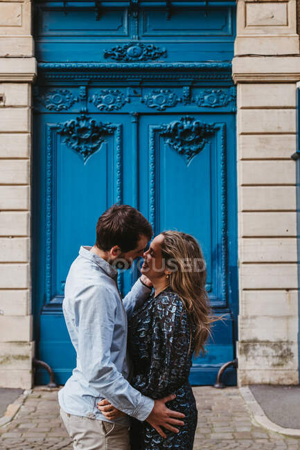 Seitenansicht eines glücklichen jungen Paares in lässiger Kleidung, das sich umarmt und küsst, während es vor einem alten Steingebäude mit blauen Türen auf der Stadtstraße steht — Stockfoto