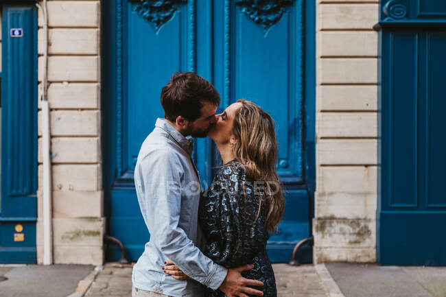 Vista lateral do jovem casal feliz em roupas casuais abraçando e beijando enquanto estava de pé contra edifício de pedra envelhecida com portas azuis na rua da cidade — Fotografia de Stock