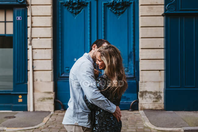 Вид збоку щасливої молодої пари в повсякденному одязі обіймає і цілує, стоячи проти старого кам'яного будинку з блакитними дверима на вулиці міста — стокове фото