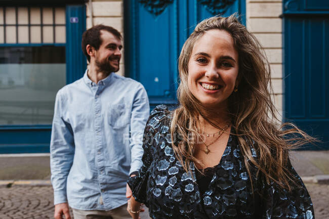 Joyeux jeune femme regardant la caméra dans une tenue élégante suivie d'un petit ami souriant marchant dans la rue de la ville avec un vieux bâtiment en arrière-plan pendant les vacances romantiques en France — Photo de stock