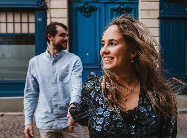 Счастливая молодая женщина в стильном наряде, за которой следует улыбающийся бойфренд, идущий по городской улице со старым зданием на заднем плане во время романтического отдыха во Франции — стоковое фото