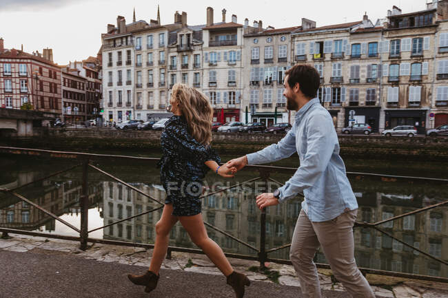 Vista lateral de feliz jovem casal afetuoso correndo com as mãos rindo em aterro de pedra perto do rio com edifícios antigos no fundo na cidade de Bayonne, na França — Fotografia de Stock