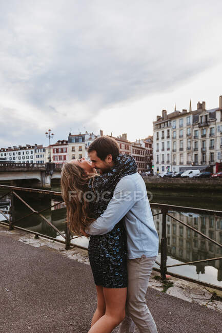 Боковой вид счастливой молодой влюбленной пары, обнимающей и целующейся, стоя на каменной набережной возле реки со старыми зданиями на заднем плане в городе Байонне во Франции — стоковое фото