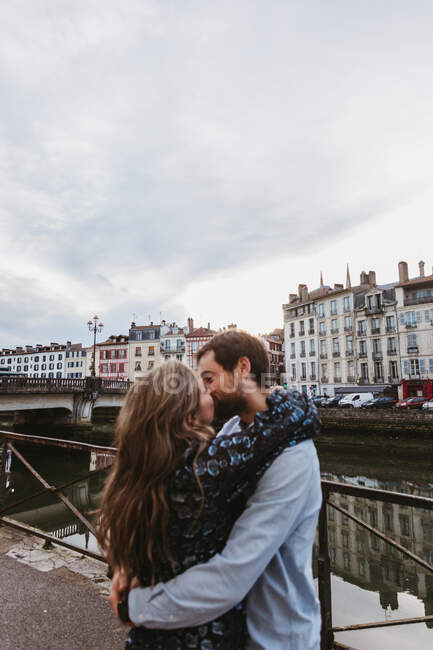 Боковой вид счастливой молодой влюбленной пары, обнимающей и целующейся, стоя на каменной набережной возле реки со старыми зданиями на заднем плане в городе Байонне во Франции — стоковое фото