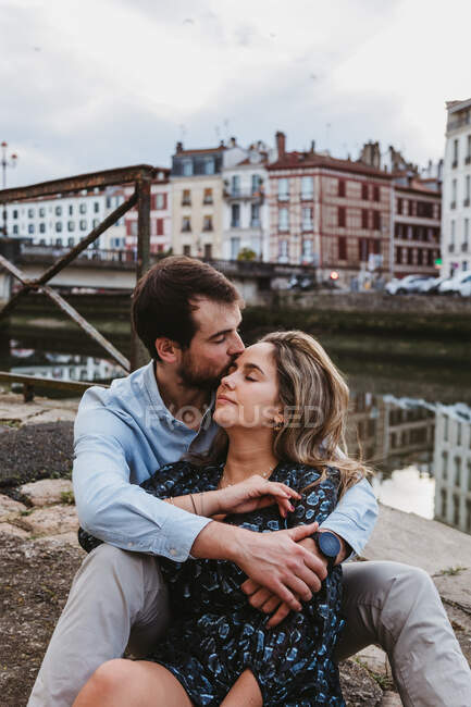 Позитивна молода пара в повсякденному одязі насолоджується романтичним побаченням, сидячи разом на кам'яній межі в місті зі старими будівлями на задньому плані — стокове фото