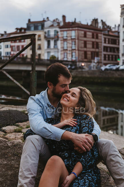 Pareja joven positiva en ropa casual disfrutando de una cita romántica mientras están sentados juntos en la frontera de piedra en la ciudad con edificios antiguos en el fondo - foto de stock