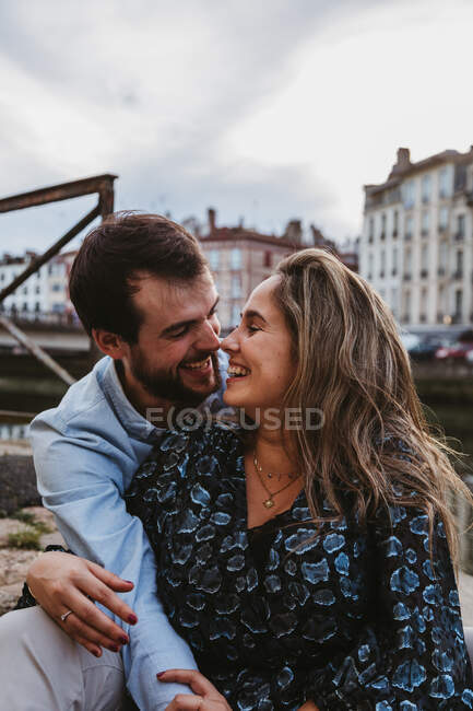 Positive young couple in casual clothes enjoying romantic date while sitting together on stone border in city with old buildings in background — Stock Photo