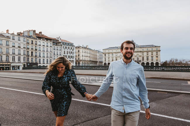 Glückliches junges romantisches Paar in stylischer Kleidung, lachend und Händchenhaltend, während es die Brücke mit historischen Gebäuden im Hintergrund während einer Stadtrundfahrt in Bayonne in Frankreich überquert — Stockfoto