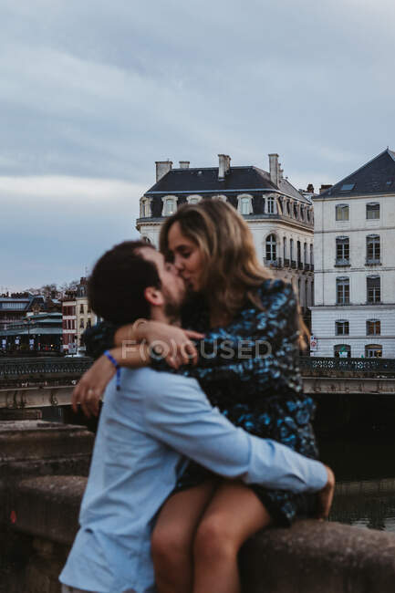 Blurred happy woman sitting on old stone fence and kissing affectionate boyfriend while spending summer evening together in Bayonne — Stock Photo