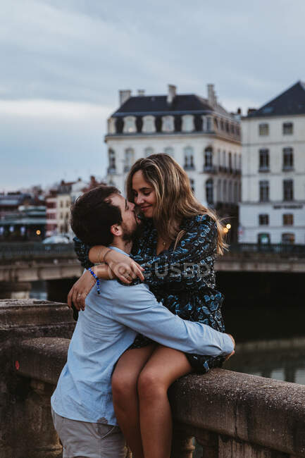 Joyeux jeune femme assise sur une vieille clôture en pierre et embrassant son affectueux petit ami tout en passant la soirée d'été ensemble à Bayonne — Photo de stock