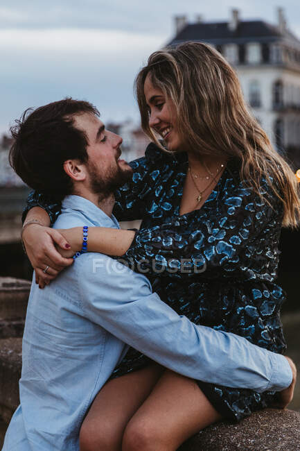 Feliz joven hembra sentada en una vieja valla de piedra y abrazando a un novio cariñoso mientras pasan la noche de verano juntos en Bayona - foto de stock