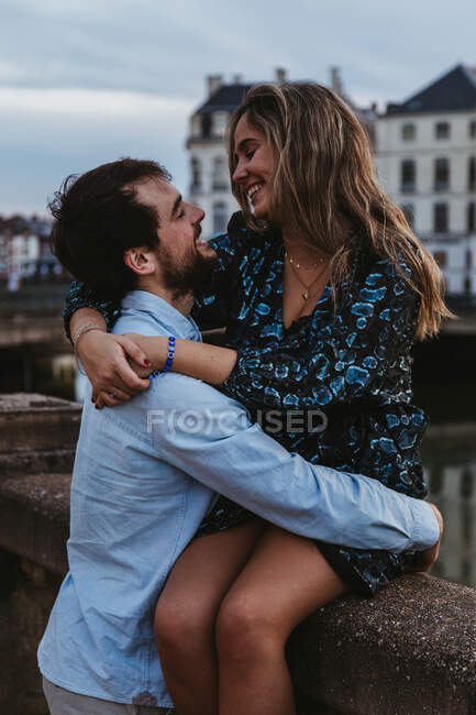 Happy young female sitting on old stone fence and embracing affectionate boyfriend while spending summer evening together in Bayonne — Stock Photo