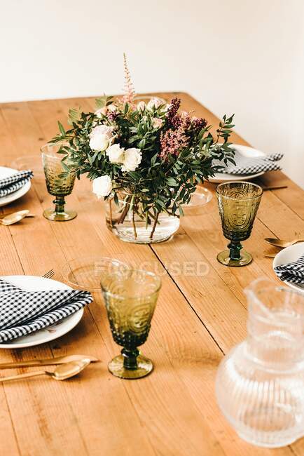 Bouquet of miscellaneous flowers and green plant twigs in vase with water on a wooden table set for a meal — Stock Photo