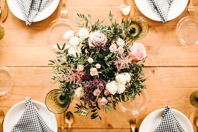 From above top view bouquet of miscellaneous flowers and green plant twigs in vase with water on a wooden table set for a meal — Stock Photo