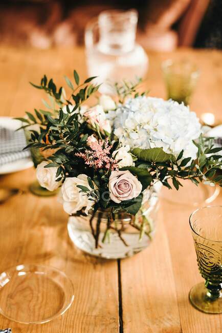 Bouquet di fiori vari e ramoscelli di piante verdi in vaso con acqua su una tavola di legno apparecchiata per un pasto — Foto stock