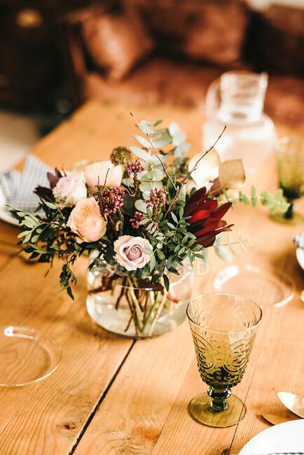 From above bouquet of miscellaneous flowers and green plant twigs in vase with water on a wooden table set for a meal — Stock Photo