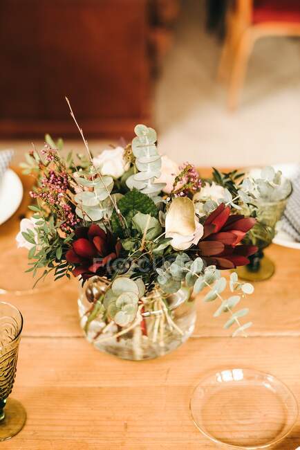 Сверху букет разноцветных цветов и зеленых цветочных веток в вазе с водой на деревянном столе, накрытом на обед — стоковое фото