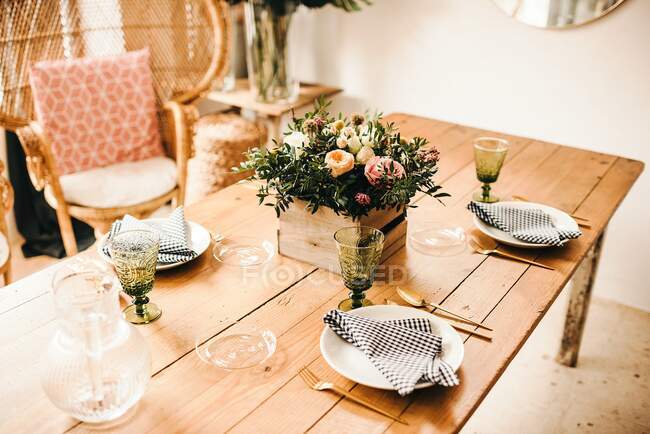 From above bouquet of miscellaneous flowers and green plant twigs in a wooden box on a timber table set for a meal with beautiful designed rattan chair on the background — Stock Photo