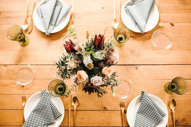 From above top view bouquet of miscellaneous flowers and green plant twigs in vase with water on a wooden table set for a meal — Stock Photo