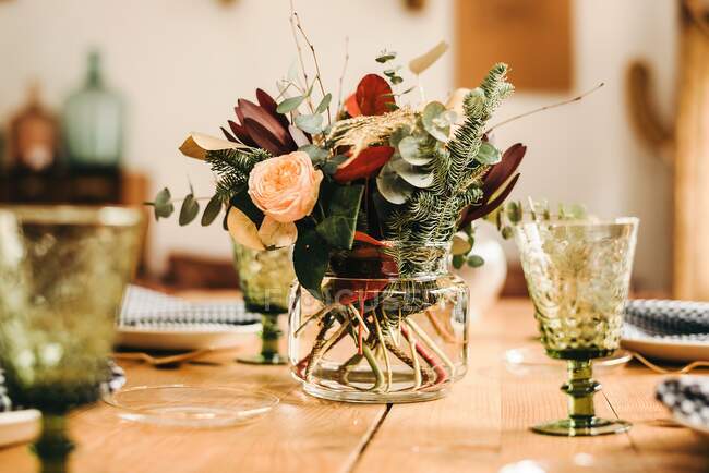 Bouquet di fiori vari e ramoscelli di piante verdi in vaso con acqua su una tavola di legno apparecchiata per un pasto — Foto stock
