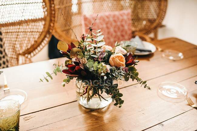 From above bouquet of miscellaneous flowers and green plant twigs in vase with water on a wooden table set for a meal with beautiful designed rattan chair on the background — Stock Photo