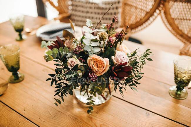 Сверху букет разноцветных цветов и зеленых цветочных веток в вазе с водой на деревянном столе, накрытом на стол для еды с красивым ротанговым стулом на заднем плане — стоковое фото