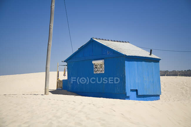 Маленький блакитний будинок з бурхливими стінами, розташований на піщаному узбережжі з блакитним небом на фоні сонячного дня — стокове фото