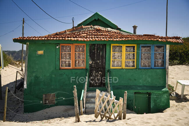 Small house with shabby green walls and colorful windows located on sandy seaside with blue sky in background in sunny day — Stock Photo
