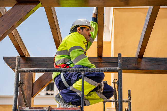 Von unten kauerte ein männlicher Techniker in Arbeitskleidung auf einem Gerüst und sah weg, als er sich auf die Installation von Solarzellen auf Holzkonstruktion vorbereitete — Stockfoto
