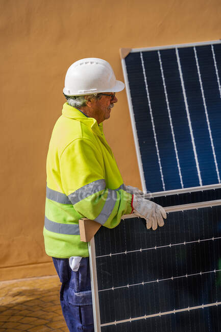 Побочный обзор опытного мужчины-техника в форме и шлеме, стоящего рядом с солнечной панелью возле желтого здания во время работы по установке системы возобновляемой энергии — стоковое фото
