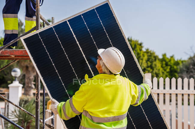 Back view of mature experienced male technician in uniform and helmet standing with solar panel near yellow building while working on installation of renewable energy system — Stock Photo
