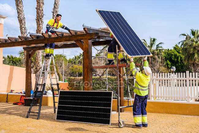 Groupe de travailleurs en uniforme et casques installant des panneaux photovoltaïques sur le toit de la construction en bois près de la maison — Photo de stock