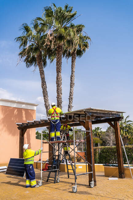 Группа работников в форме и шлемах, устанавливающих фотоэлектрические панели на крыше деревянной конструкции рядом с домом — стоковое фото