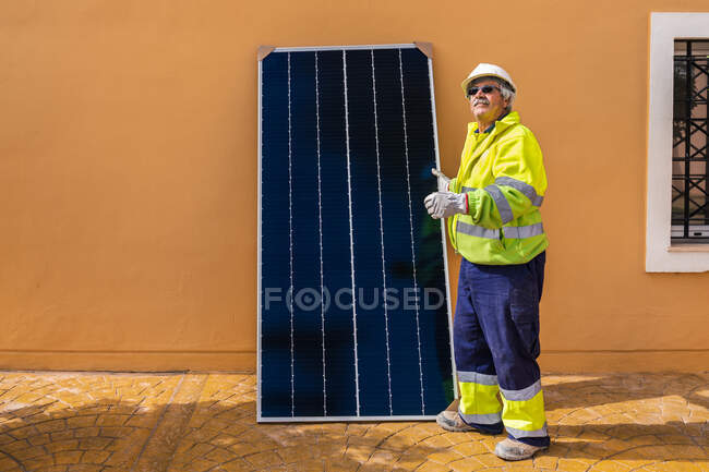 Side view of mature experienced male technician in uniform and helmet standing with solar panel near yellow building while working on installation of renewable energy system — Stock Photo