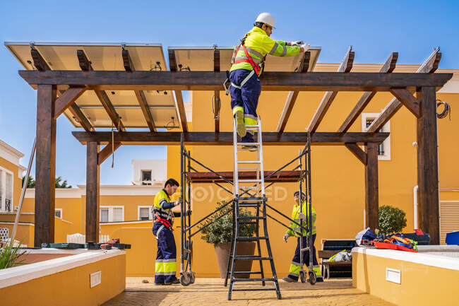 Gruppo di lavoratori in uniforme e caschi installazione pannelli fotovoltaici sul tetto di costruzione in legno vicino casa — Foto stock
