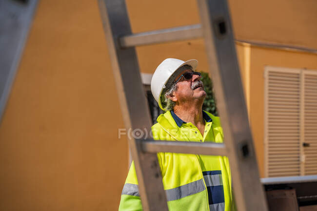 Trabalhador de cabelos grisalhos qualificados positivos em capacete uniforme e protetor olhando para longe enquanto estava atrás da construção de metal contra a parede do edifício em dia ensolarado — Fotografia de Stock