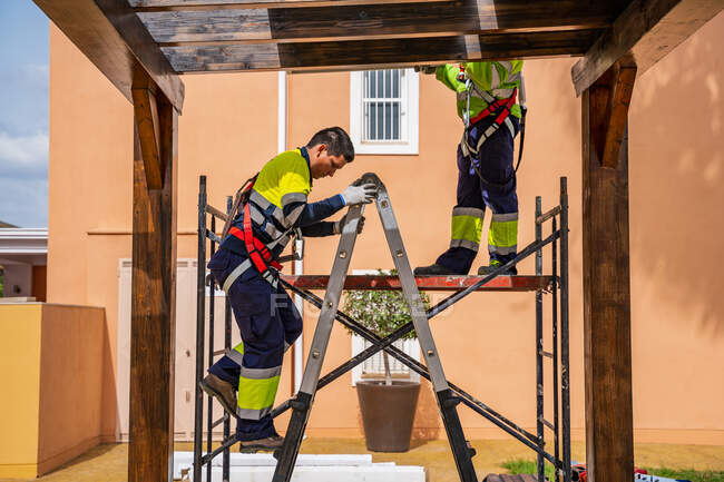 Groupe de travailleurs en uniforme et casques installant des panneaux photovoltaïques sur le toit de la construction en bois près de la maison — Photo de stock