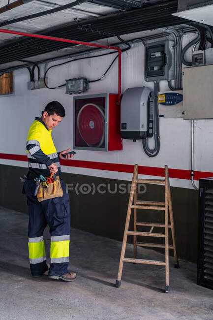 Vista laterale di esperto ingegnere uomo in uniforme utilizzando il telefono cellulare durante l'esame di apparecchiature elettriche in un edificio moderno — Foto stock