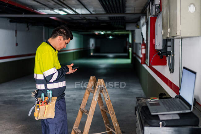 Vista lateral del ingeniero experto hombre en uniforme utilizando el teléfono móvil mientras se examina el equipo eléctrico en el edificio moderno - foto de stock