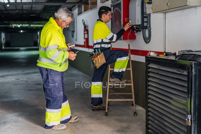 Seitenansicht einer Gruppe professioneller männlicher Techniker mit elektrischen Werkzeugen, die Ausrüstung reparieren und überprüfen, während sie im Gebäude arbeiten — Stockfoto