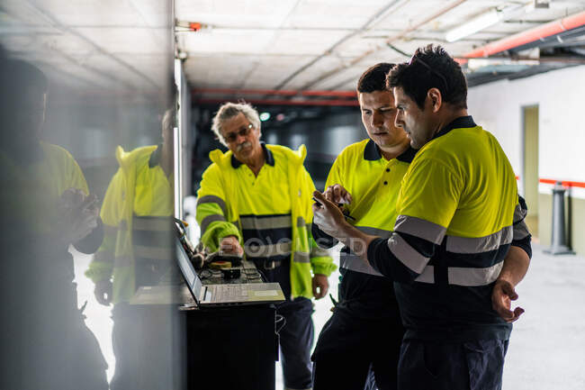 Grupo de engenheiros do sexo masculino qualificados em uniforme usando gadgets enquanto examina equipamentos elétricos no edifício moderno — Fotografia de Stock