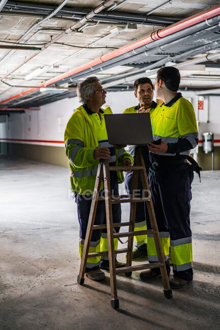 Grupo de engenheiros do sexo masculino qualificados em uniforme usando gadgets enquanto examina equipamentos elétricos no edifício moderno — Fotografia de Stock
